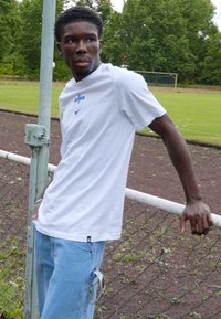 Weißes Nike-T-Shirt mit blauem Logo und Finnland-Symbol, kombiniert mit hellblauen Jeans. Steht an einem Maschendrahtzaun auf einem Grasfeld.