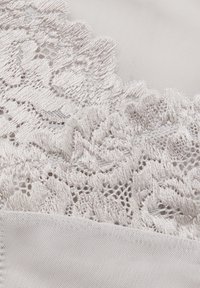 Lätt tyg med intrikat vit blommig spetskant. Spetsen har en scalloped kant och texturerade detaljer, vilket kontrasterar mot en slät bakgrund.
