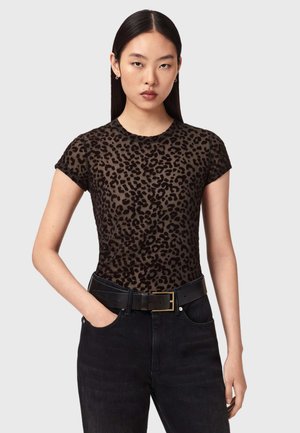 Ung kvinde med glat mørkt hår iført en kortærmet top med leopardprint, sort bælte og sorte bukser, hænderne i lommerne, neutral baggrund.