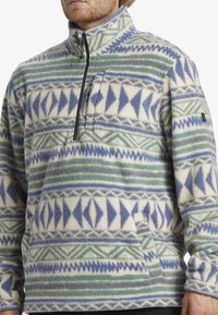Pullover fleece jakke med geometriske mønstre i blå, grøn og lilla på en cremefarvet baggrund. Har en halvlukning og en frontlomme.