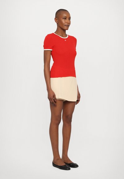 Haut en tricot rouge à côtes avec des manches courtes et un ourlet blanc, assorti à une mini-jupe beige extensible. Le mannequin porte des chaussures plates noires.