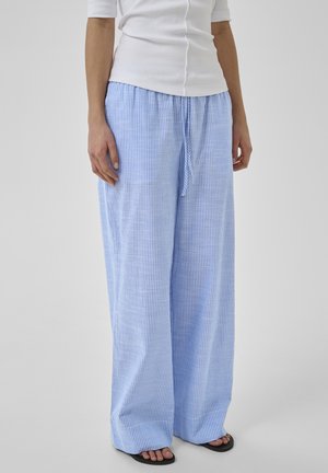 Nő világoskék és fehér, függőleges csíkos, bőszárú nadrágban, húzózsinóros derékrésszel, fehér testhezálló felsővel és fekete szandállal párosítva.