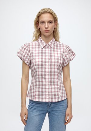 Femme blonde portant une chemise blanche à manches courtes avec un motif à carreaux rouges et un jean bleu, debout devant un fond blanc uni.