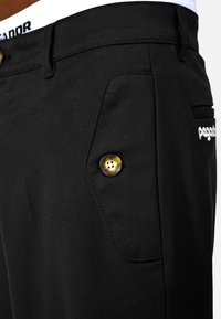 Melnas bikses, izgatavotas no struktūrēta auduma, ar izteiktu zelta pogu un izšūtu logotipu. Iespējama sānu kabata ar elegantu dizainu.