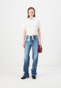 Chemise blanche à manches courtes avec un design en œillets, nouée à la taille. Jean bleu rayé, coupe droite, associé à un petit sac à main rouge.
