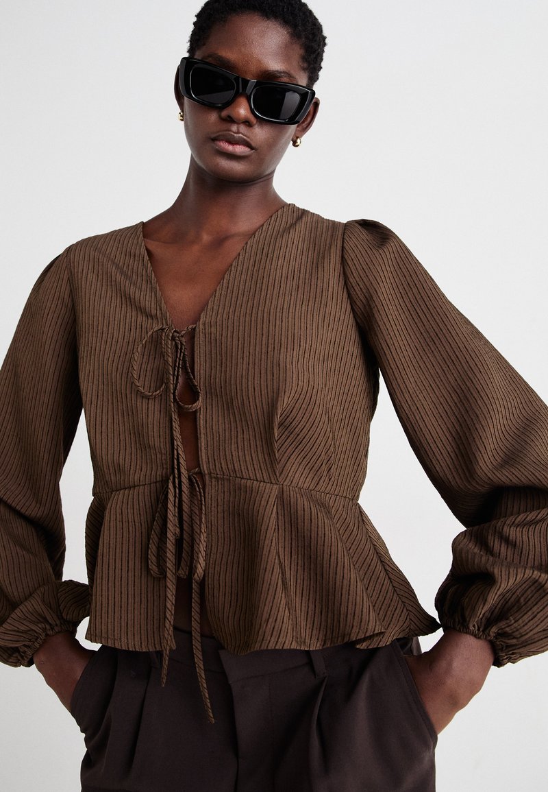 Brązowa, prążkowana bluzka z dekoltem w kształcie litery V, cinzowanym wcięciem w talii i bufiastymi rękawami. Posiada zapięcie na kokardkę z przodu oraz pionowy wzór w paski.