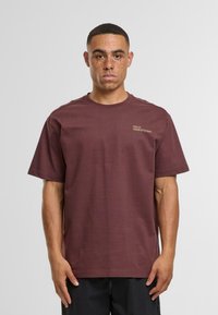 Mladý muž s krátkými vlasy a pihami, oblečený v tmavě červeném tričku s malým žlutým textem na levé hrudi, stojící na neutrálním světlém pozadí.