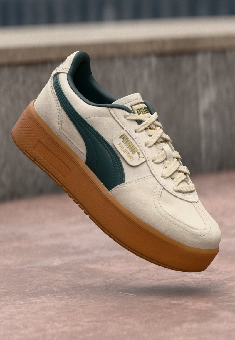 Beige suede sneakers with a dark green side accent, textured gum sole, and gold logo detail on the tongue. Lightweight design with laces.