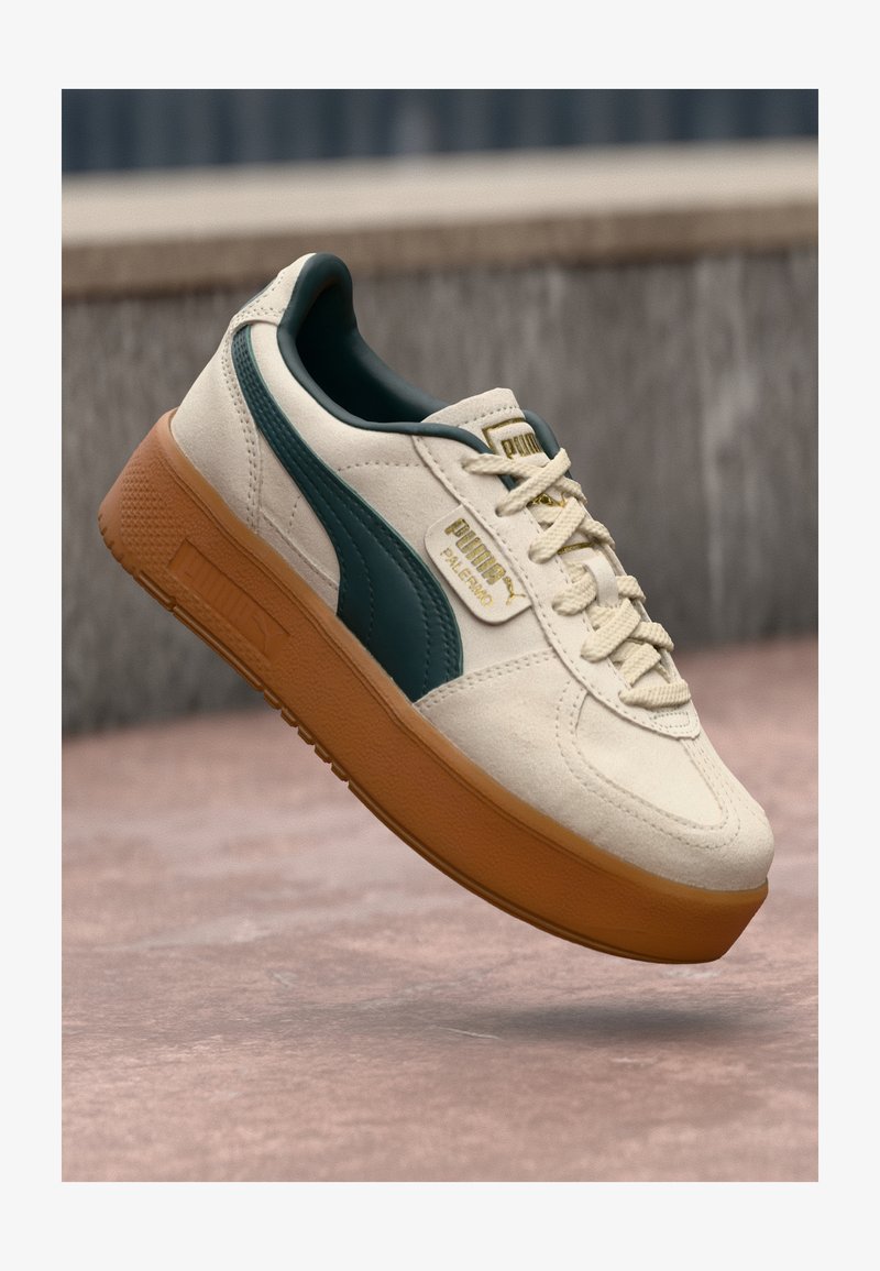 Beige suede sneakers with a dark green side accent, textured gum sole, and gold logo detail on the tongue. Lightweight design with laces.