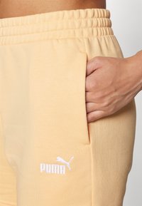 Ljus persikofärgade joggerbyxor har en elastisk midja, sidoficka och en liten vit Puma-logotyp nära fållen. Slät textur, bomullsblandat material.