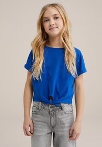 Blå kortærmet t-shirt med en knyttet frontdetalje, lavet af glat stof, parret med lysegrå højtaljede jeans.