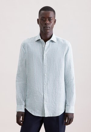 Mand iført en langærmet skjorte med lodrette grønne og hvide striber og mørke bukser, står foran en ensfarvet baggrund.