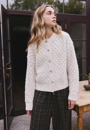 Junge Frau mit welligem blondem Haar, die eine cremefarbene Strickjacke und dunkel karierte Hose trägt, steht nahe einer offenen Tür und einem Holztisch.