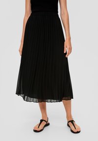 Kvinna i svart ärmlös topp, svart plisserad midikjol och svarta sandaletter i thong-stil som står mot en enfärgad bakgrund.