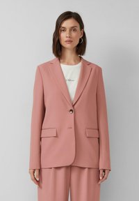 Vrouw in een mauve colbert met een enkele knoop en bijpassende broek over een wit shirt met kleine tekst.