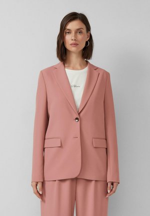 Frau in einem mauvefarbenen Blazer mit einem einzelnen Knopf und passenden Hosen über einem weißen Hemd mit kleiner Schrift.