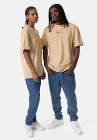 Δύο άτομα φορούν μπεζ φαρδιά μπλουζάκια με μια μαύρη λογότυπο στο στήθος, συνδυασμένα με μπλε τζιν παντελόνια και λευκά αθλητικά παπούτσια.