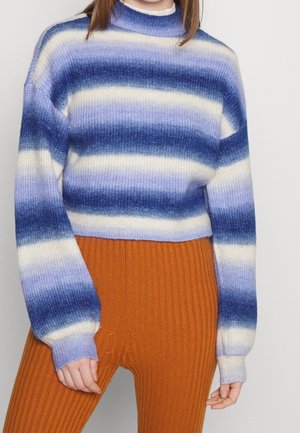 Personne portant un pull en maille rayé bleu, blanc et violet et un pantalon côtelé orange taille haute sur un fond uni.