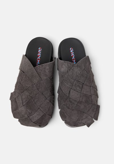 Gray suede slip-on slippers with a woven texture and open toe. Black insoles feature colorful branding. Soft material and casual design.