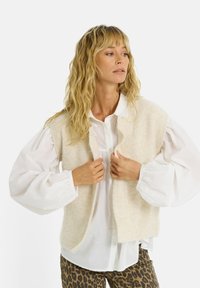Mujer con cabello rubio ondulado, vestida con una camisa blanca de mangas abullonadas, un chaleco de punto sin mangas color crema y pantalones de estampado de leopardo, ajustándose el chaleco.