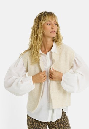 Mujer con cabello rubio ondulado, vestida con una camisa blanca de mangas abullonadas, un chaleco de punto sin mangas color crema y pantalones de estampado de leopardo, ajustándose el chaleco.