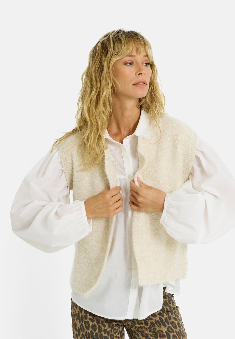 Mujer con cabello rubio ondulado, vestida con una camisa blanca de mangas abullonadas, un chaleco de punto sin mangas color crema y pantalones de estampado de leopardo, ajustándose el chaleco.