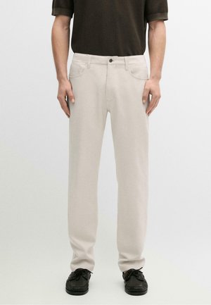 Mann trägt beige gerade Hose, schwarze Schuhe und ein dunkles kurzärmeliges Hemd und steht vor einem schlichten Hintergrund.