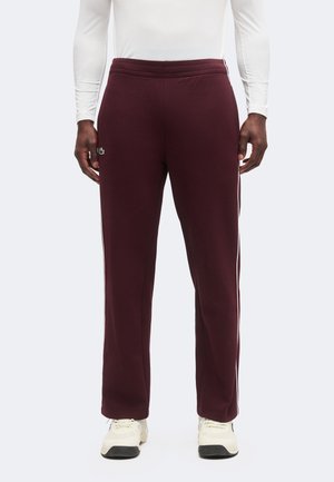 Homme portant un jogging bordeaux foncé avec des bandes blanches sur les côtés, des baskets blanches et un t-shirt blanc ajusté à manches longues, debout face à l'avant.