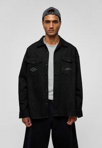 Zwarte button-up shirt met twee voorkruiszakken en een diamanten patch, gemaakt van textuurstof, gedragen over een grijze T-shirt.