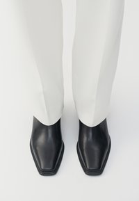 Crne kožne gležnjače s šiljastim nosom i niskim složenim potplatom, uparene s bijelim hlačama širokih nogavica. Glatka tekstura, elegantan dizajn.
