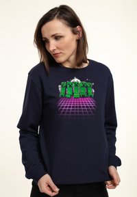 Felpa blu navy con una grafica colorata di personaggi verdi pixelati su uno sfondo a griglia retro rosa. Tessuto morbido, vestibilità casual.