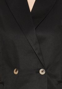 Femme portant un blazer noir sur mesure avec de larges revers et deux boutons beiges fermant le devant.