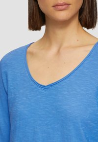 Haut à manches longues bleu en V, en tissu texturé. Fini lisse, avec une coupe décontractée et un motif chiné subtil.