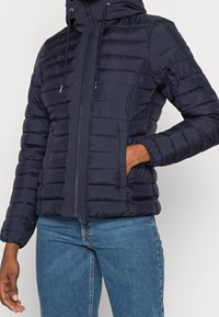 Person in einer dunkelblauen Steppjacke mit Kapuze und blauen Jeans, Hand in der Jackentasche, stehend vor einem schlichten Hintergrund.