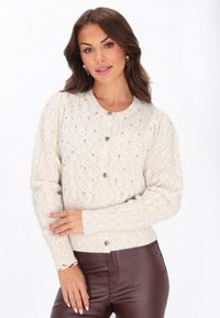 Könnyű bézs kötött kardigán texturált mintával, kerek nyakkivágással és dekoratív gombokkal. Elöl gyöngyös díszítésekkel rendelkezik.