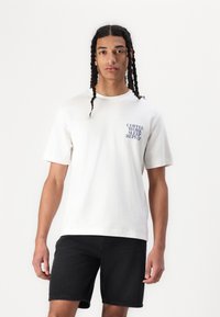 Kurzärmliges weißes Baumwoll-T-Shirt mit blauem Text „KAFFEE ARBEITEN SCHLAFEN WIDERHOLEN“ auf der linken Brust; kombiniert mit schwarzen Jeansshorts.