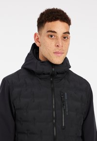 Schwarze Daunenjacke mit hohem Kragen, Reißverschluss vorne und einem Kontrastpaneel. Verfügt über eine Reißverschlusstasche auf der Brust und eine glatte Textur.