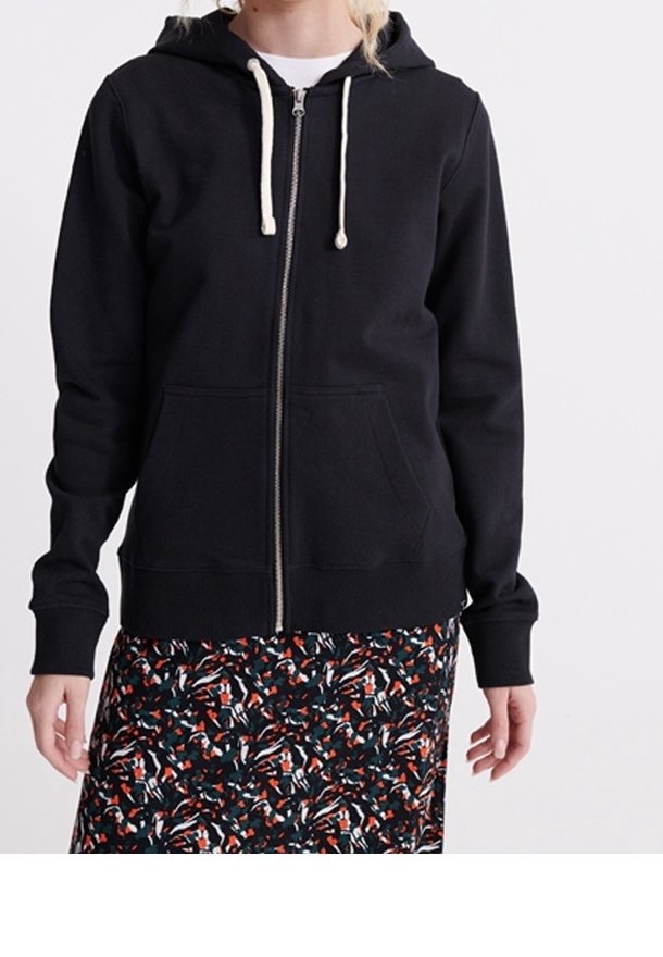 Personne portant un sweat à capuche noir zippé avec des cordons blancs et une jupe à motifs noir, blanc et rouge.