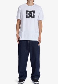 Osoba v bílém tričku s krátkým rukávem s černým logem DC, volných tmavě modrých jeansách a černých teniskách stojící na bílém pozadí.