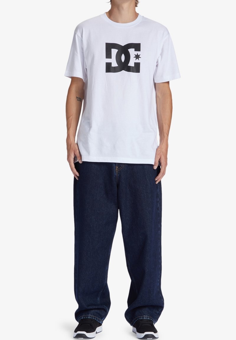 Osoba v bílém tričku s krátkým rukávem s černým logem DC, volných tmavě modrých jeansách a černých teniskách stojící na bílém pozadí.