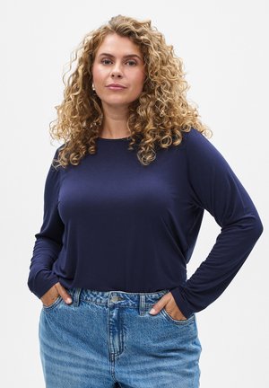 Long-sleeve navy shirt made of soft fabric, paired with light blue high-waisted denim jeans. Curly hair frames the face.