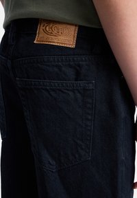 Tmavé džíny z denimu s hnědým koženým logem na pásku, s viditelným šitím a detailem zadní kapsy. Měkká textura.