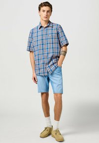 Camicetta a maniche corte, a quadretti, blu e rosa, con taschino sul petto, abbinata a pantaloni corti di denim chiaro e scarpe in suede beige. Calzini bianchi visibili.