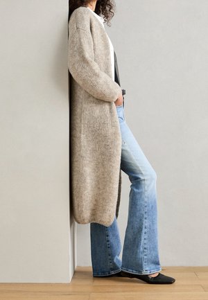 Osoba opierająca się o ścianę, ubrana w beżowy długi kardigan, białą koszulę, rozkloszowane niebieskie dżinsy i czarne spiczaste baleriny.