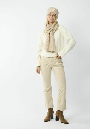 Beige gebreide beanie en sjaal gecombineerd met een witte geribbelde sweater, beige broek en bruine enkellaarsjes met een blokhak.
