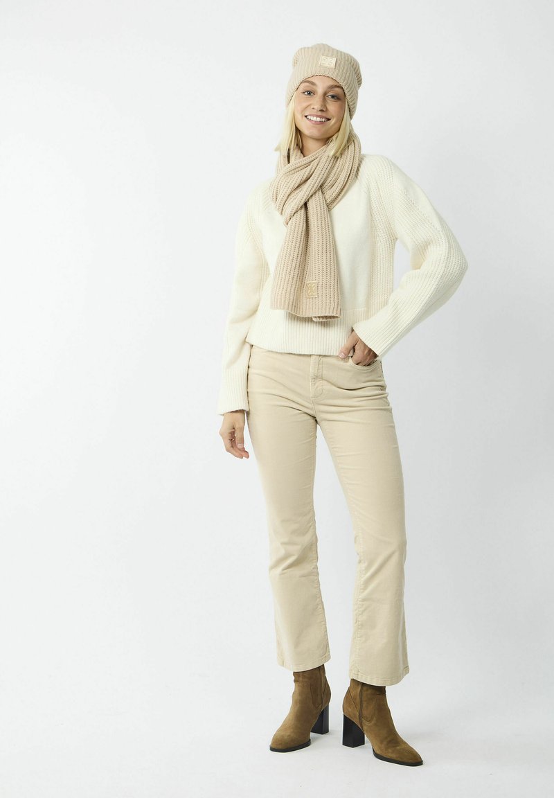 Beige gestrickte Mütze und Schal kombiniert mit einem weißen Rippenpullover, beigen Hosen und braunen Stiefeletten mit Blockabsatz.