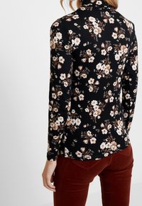 Långärmad svart topp med ett blommönster i vita och bruna toner, med hög hals och figurnära design, kombinerad med röda kordbyxor.