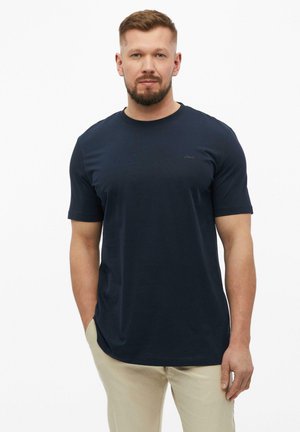 Mężczyzna z krótkimi jasnobrązowymi włosami i brodą, ubrany w granatowy t-shirt z krótkim rękawem i beżowe spodnie, stojący na białym tle.