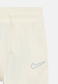 Kremkrāsas treniņbikses ar ribotu jostasvietu, auklu un pelēku izšūtu Nike logo kreisajā pusē. Mīksts, teksturēts audums.