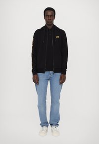 Zwarte zip-up hoodie met gele "EA7" logo op de mouw, gelaagd over een polo shirt, gecombineerd met lichtblauwe jeans en witte sneakers.